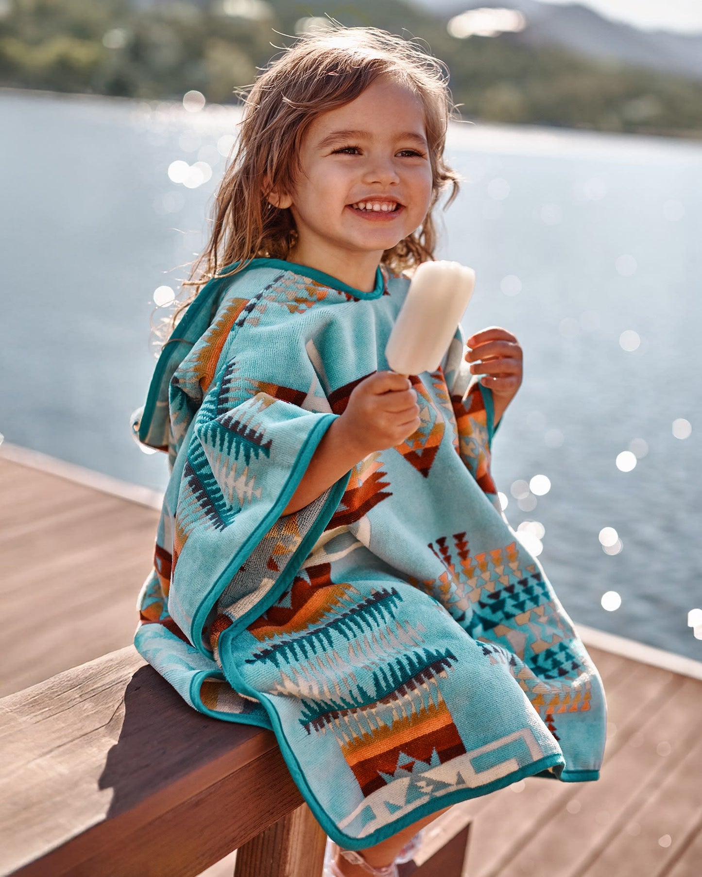 Child wearing a patterned poncho by a lake holding a popsicle.